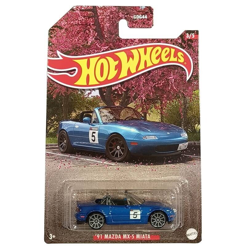 Hot Wheels 1:64 Walmart Exclusive Theme JDM toy car in packaging, featuring a blue Mazda MX-5 Miata with racing decals, shown with a real car beneath pink flowering trees. Brand: Hot Wheels.