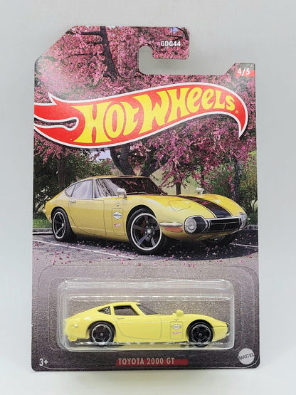 A yellow Hot Wheels 1:64 Toyota 2000 GT from the Walmart Exclusive Theme JDM set features packaging with pink blooming trees, car illustration, and a large Hot Wheels logo.