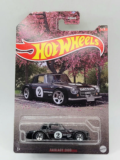 Hot Wheels 1:64 Walmart Exclusive Theme JDM toy car, featuring a black Datsun Fairlady 2000 with number 2 on the side. The packaging shows the car parked under blooming trees. Brand: Hot Wheels.